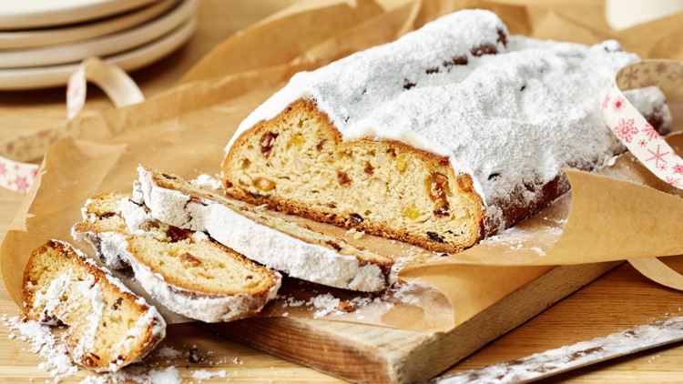 Ein aufgeschnittener Stollen mit Puderzucker liegt auf Backpapier, daneben ein Messer und ein Band mit weihnachtlichem Muster.