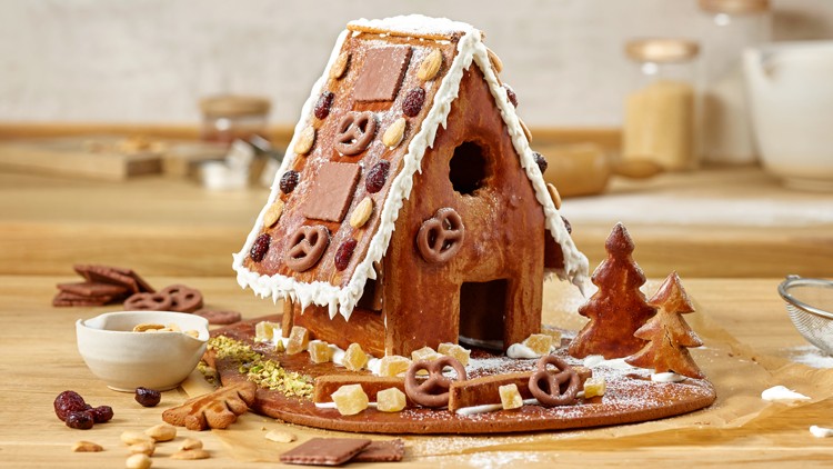 Lebkuchenhaus mit Zuckerguss, Schokoladendekoration und kleinen Gebäckbäumen auf einem Holztisch, daneben Backzutaten.