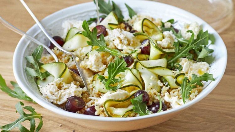 Ein Teller mit Rucola, Zucchinistreifen, Trauben, zerbröckeltem Käse und Bulgur auf einem Holztisch, daneben ein Servierlöffel.