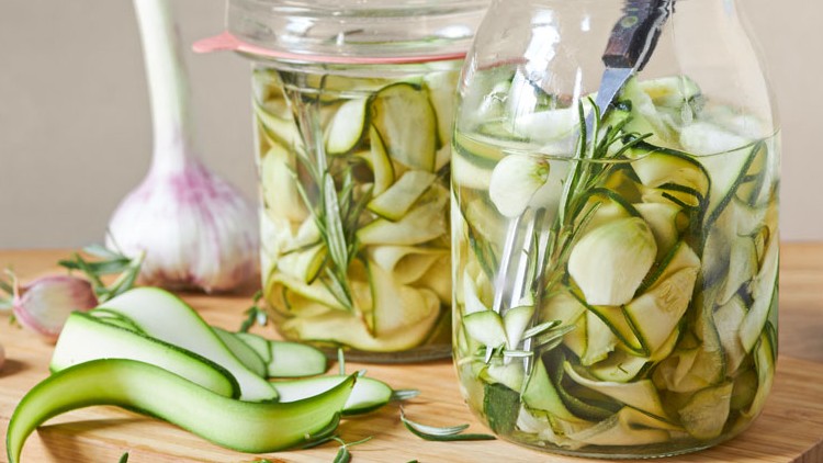 Zwei Gläser mit eingelegten Zucchinistreifen und Rosmarinzweigen, daneben frischer Knoblauch und ein Sparschäler auf Holzbrett.