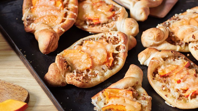 Kleine herzhafte Teigschiffchen mit Kürbisscheiben und Käse auf einem Backblech, daneben ein aufgeschnittener Kürbis auf Holzbrett.