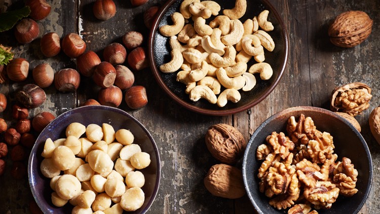 Verschiedene Nüsse wie Haselnüsse, Macadamianüsse, Cashewnüsse und Walnüsse liegen teils in Schalen, teils lose auf Holztisch.