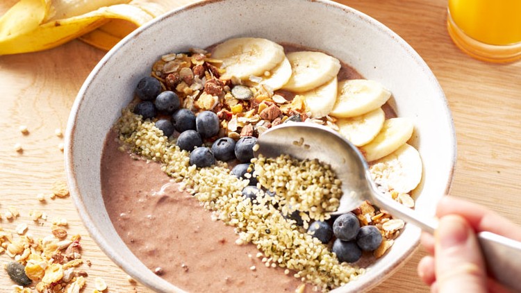 Eine helle Schale mit Bananenscheiben, Blaubeeren, Müsli und Samen darauf, daneben ein Glas Orangensaft und eine Banane.