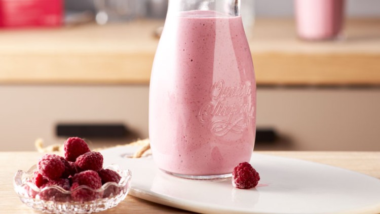 Glasflasche mit rosa Himbeer-Smoothie auf weißem Teller, daneben eine kleine Schale mit frischen Himbeeren.