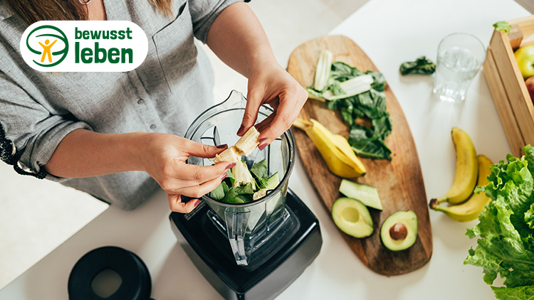 Hände geben Stücke einer Banane in einen Mixer mit Spinat und Gurken, daneben liegen Avocado, Salat und weitere Zutaten.