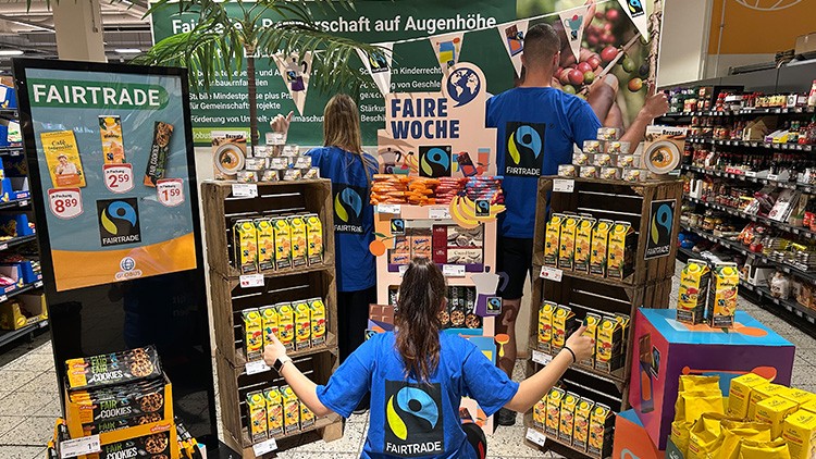 Drei Personen in FAIRTRADE-Shirts stehen vor einem Verkaufsstand mit Fairtrade-Produkten, Text „FAIRE WOCHE“ und Logo sichtbar.