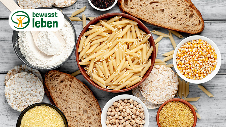 Eine Schale mit Penne, Mehl mit Holzlöffel, Maiskörner, Reiswaffeln und Brotscheiben auf hellem Holztisch, Text: bewusst leben.