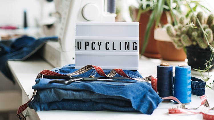 Leuchtbox mit dem Wort „UPCYCLING“ neben Stoff, Schere, Maßband und Nähgarn auf einem Tisch, im Hintergrund Pflanzen.