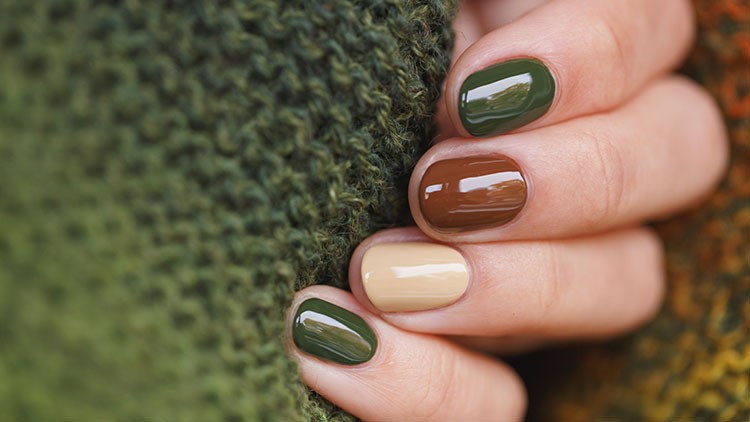 Eine Hand mit lackierten Nägeln in Grün, Braun und Beige hält gestricktes Gewebe in verschiedenen Grüntönen.