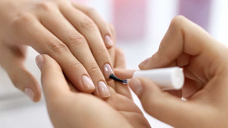 Eine Hand lackiert die Fingernägel einer anderen Hand mit einem transparenten Pinsel.