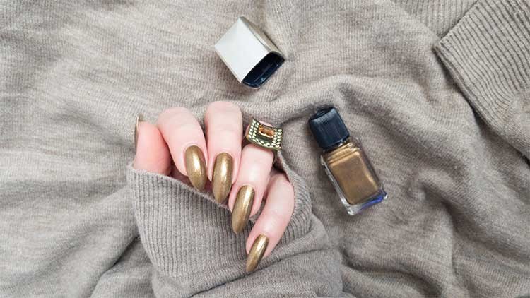 Eine Hand mit goldfarben lackierten Nägeln hält ein beigees Oberteil, daneben liegen Nagellackflasche und geöffneter Deckel.