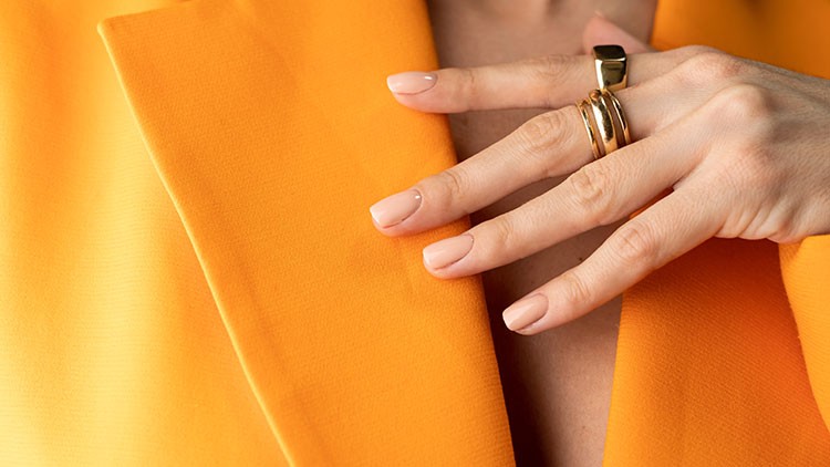 Hand mit dezent lackierten Nägeln und goldenen Ringen berührt den Kragen eines orangefarbenen Blazers.