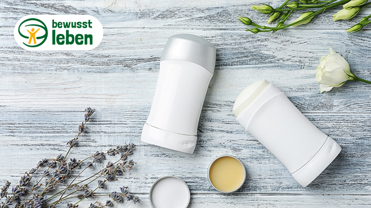 Zwei weiße Deoroller, ein offener Tiegel mit Lippenbalsam, Lavendel und eine weiße Blume auf einer hellen Holzfläche, oben links das Logo bewusst leben.
