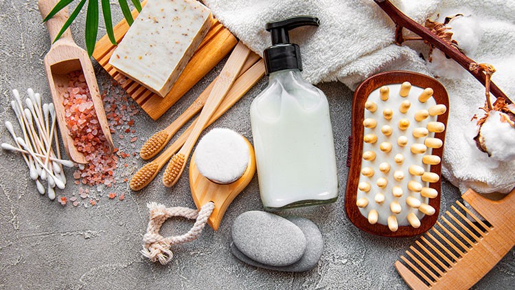 Verschiedene Badutensilien wie Seife, Creme, Massagebürste, Wattepads, Zahnbürsten und Baumwolle auf grauer Fläche.