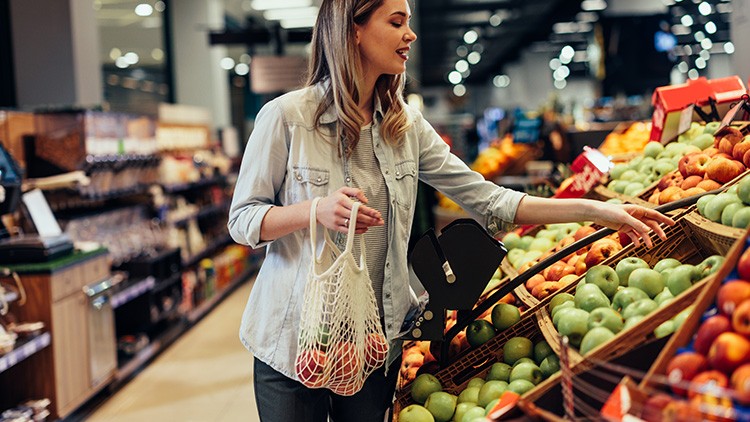 Person in hellem Hemd greift zu grünem Apfel, hält Netzbeutel mit Obst, im Hintergrund Obst- und Gemüseabteilung.