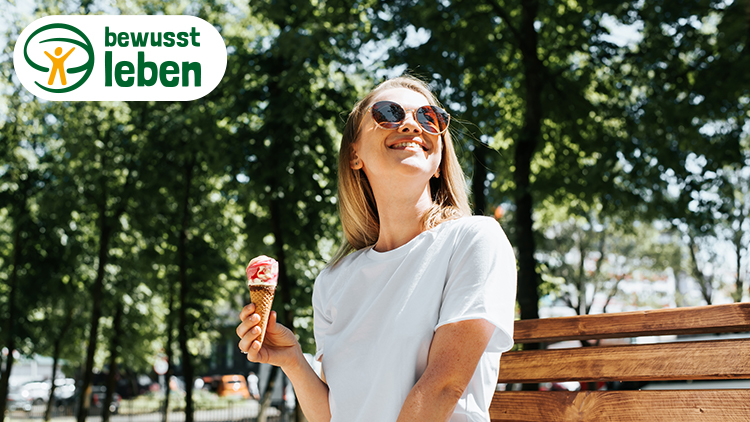 Person in weißem T-Shirt hält ein Eis mit Erdbeer- und Vanillecreme, im Hintergrund Bäume und Schriftzug „bewusst leben“.