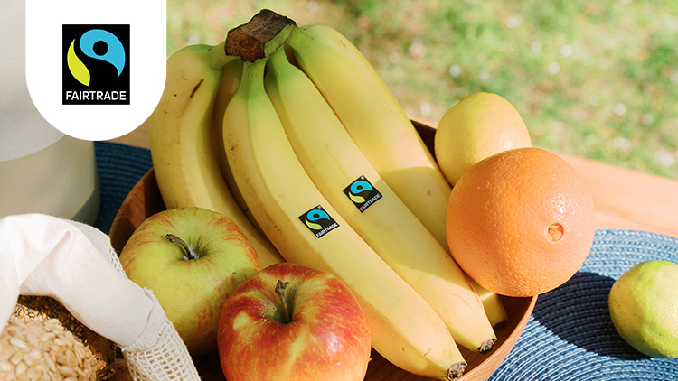 Bananen mit Fairtrade-Siegel, Äpfel, eine Orange und eine Limette liegen in einer Holzschale auf einem blauen Platzdeckchen.