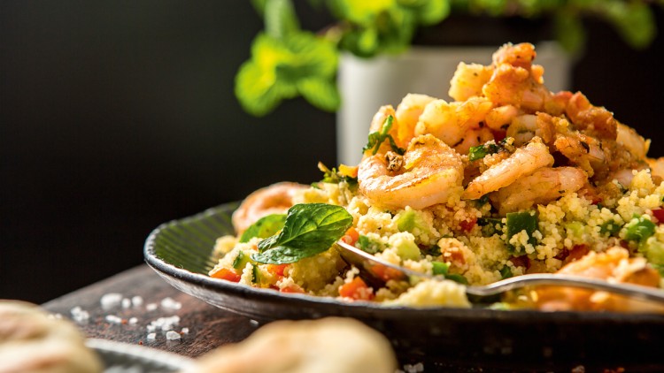 Ein Teller mit gebratenen Garnelen auf Couscous mit Gemüse und Minzblättern, im Hintergrund grüne Pflanze, unscharf.