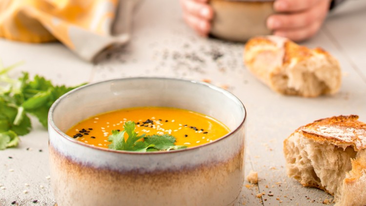 Keramische Schalen mit Kürbissuppe, dekoriert mit Koriander und Sesam, auf hellem Holztisch mit Brotstücken und Kräutern.