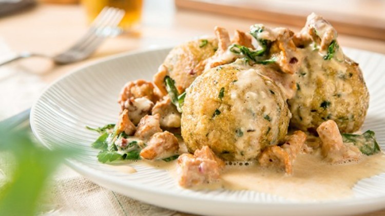 Zwei Semmelknödel in Sahnesoße mit Pilzen und Kräutern auf einem weißen Teller, im Hintergrund ein Glas Getränk.