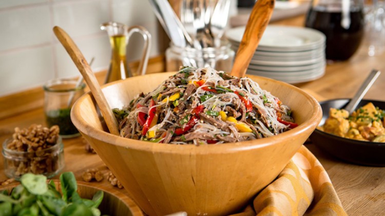 Holzschale mit Glasnudelsalat, rotem Paprika, Mais und Kräutern, daneben Walnüsse, Ölflasche, Tellerstapel und Besteck.