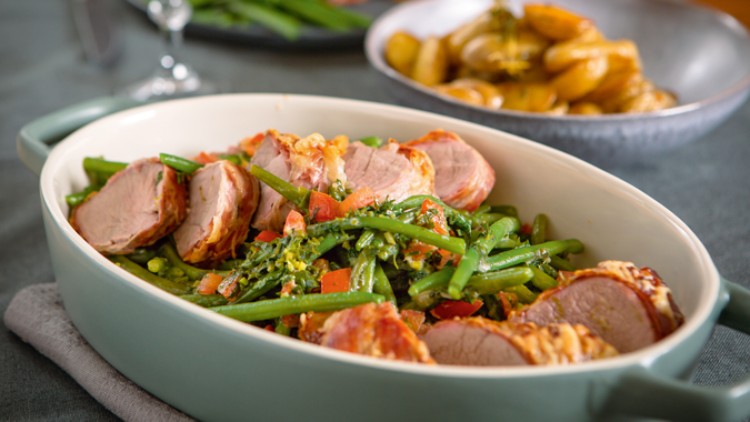In einer grünen Auflaufform liegen Schweinefiletscheiben mit grünen Bohnen, Brokkoli und Tomatenstückchen auf einem Tisch.