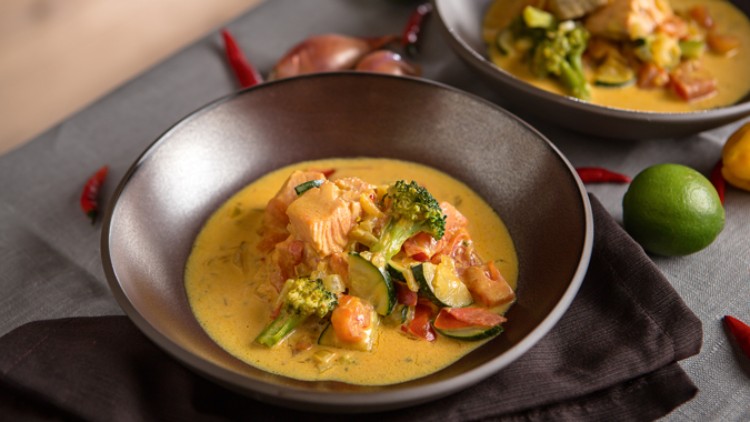 Eine Schale mit gelbem Curry, Brokkoli, Zucchini, Tomaten und Lachs steht auf einem dunklen Tuch, daneben liegen Chili und Limette.