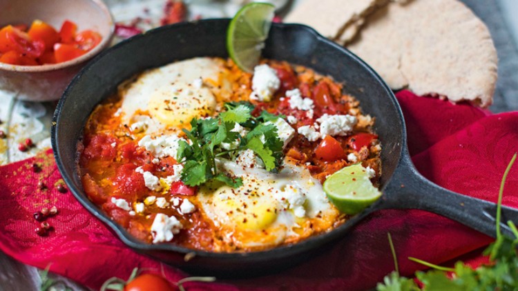 Eine schwarze Pfanne mit Shakshuka, dekoriert mit frischen Kräutern, Limettenscheiben und zerbröseltem Käse, steht auf rotem Tuch.