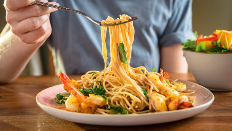 Eine Person hebt mit einer Gabel Spaghetti mit Garnelen und Spinat von einem Teller, daneben steht ein bunter Salat in einer Schale.