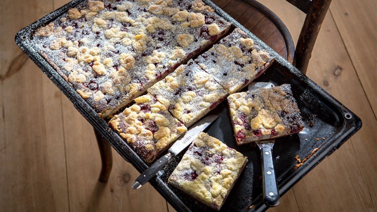 Ein Blech mit geschnittenem Streuselkuchen, teils mit Kirschen gefüllt, bestäubt mit Puderzucker, auf einem Holzstuhl.
