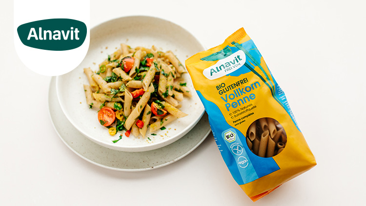 Eine geöffnete Packung Alnavit Bio Glutenfrei Vollkorn Penne neben einem Teller mit Penne, Tomaten und Kräutern.