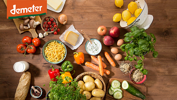 Holztisch mit Gemüse, Kräutern, Kartoffeln, Nudeln, einem Baguette, Eiern, Milch, Reis, Käse, Dip und dem Demeter-Logo oben links.