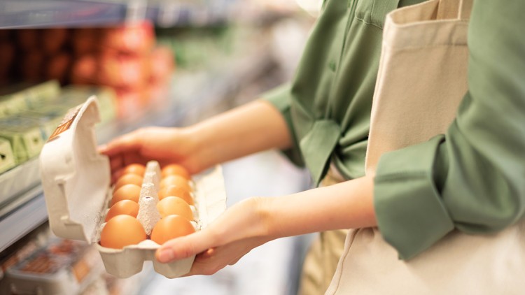 Eine Person hält einen geöffneten Karton mit zehn braunen Eiern vor einem Supermarktregal.