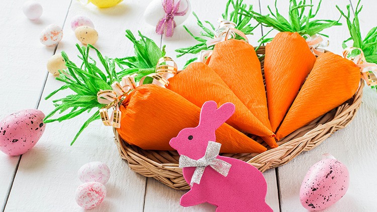 Orange Verpackungen in Karottenform liegen in einem Korb auf weißem Tisch, daneben rosa Dekohase und bunte Ostereier.