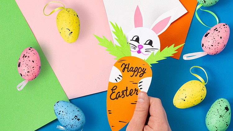Eine Hand hält eine orangefarbene Papierkarotte mit Text „Happy Easter“, umgeben von bunten Ostereiern auf farbigem Hintergrund.