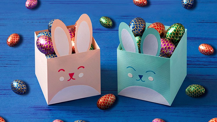 Zwei Osterboxen in Form von Hasen, gefüllt mit bunt glänzenden Schoko-Eiern, liegen auf einem blauen Holztisch.