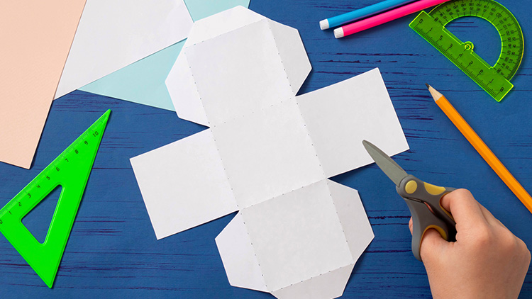 Person schneidet eine weiße Papierform aus, daneben Lineal, Geodreieck, Bleistift und farbige Stifte auf blauem Hintergrund.