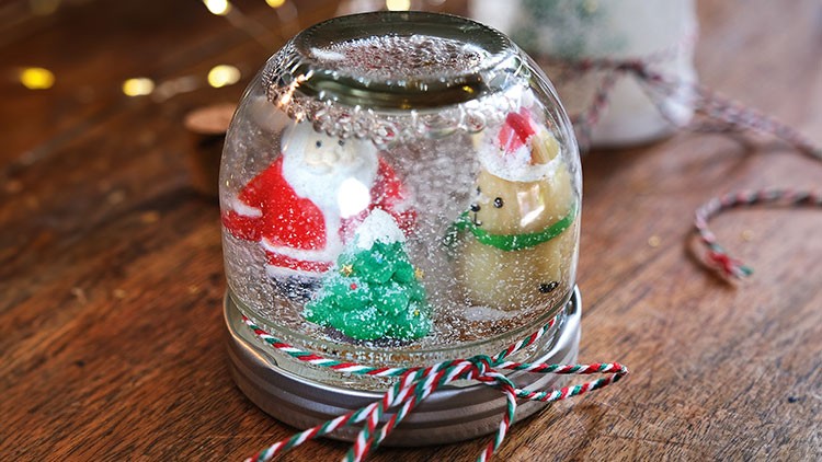 Eine Schneekugel mit Weihnachtsmann, Tannenbaum und Hund auf Holzuntergrund, verziert mit rot-weiß-grüner Kordel.