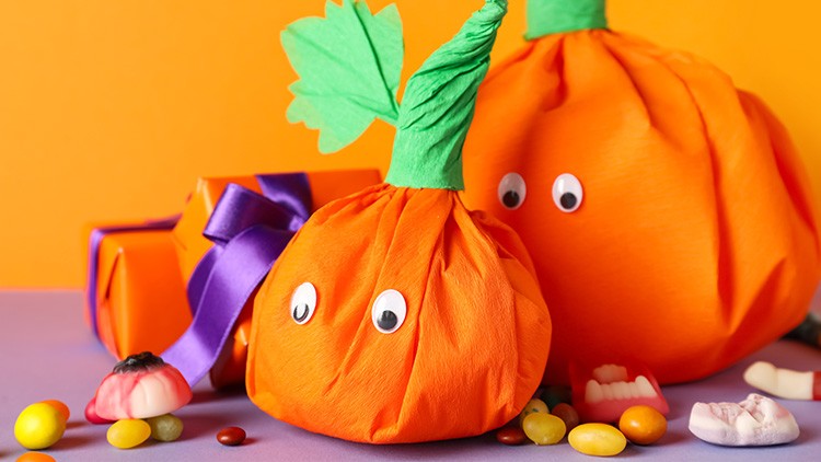 Zwei orange Verpackungen in Kürbisform mit Augen, grünem Papier oben und bunten Süßigkeiten auf lila und orangefarbenem Hintergrund.