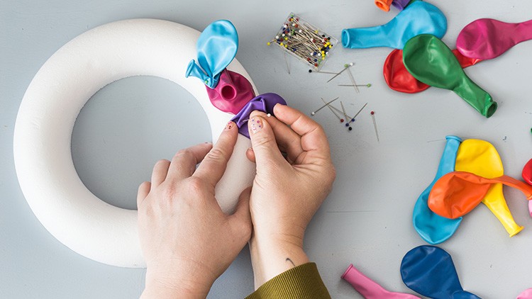 Hände befestigen bunte Luftballons mit Stecknadeln an einen weißen Styroporkranz, daneben liegen weitere Luftballons und Nadeln.