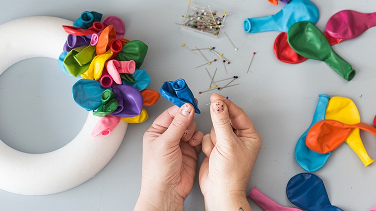 Hände stecken bunte Luftballons mit Stecknadeln auf einen weißen Styroporkranz, weitere Ballons und Nadeln liegen daneben.