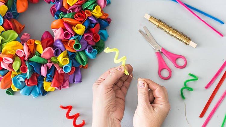 Zwei Hände formen gelben Pfeifenreiniger, daneben bunter Luftballonkranz, goldener Draht, Schere und bunte Pfeifenreiniger.