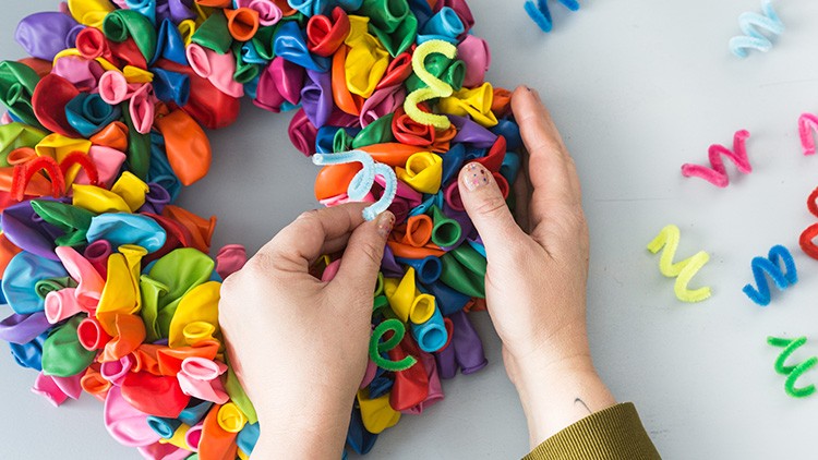 Zwei Hände formen einen Kranz aus bunten Luftballons und biegen farbige Pfeifenreiniger zu spiralförmigen Dekorationen.
