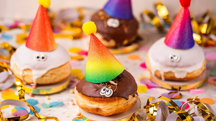 Fünf Berliner mit bunten Partyhütchen, Augen aus Zuckerguss und Konfetti auf einem Tisch mit Luftschlangen.