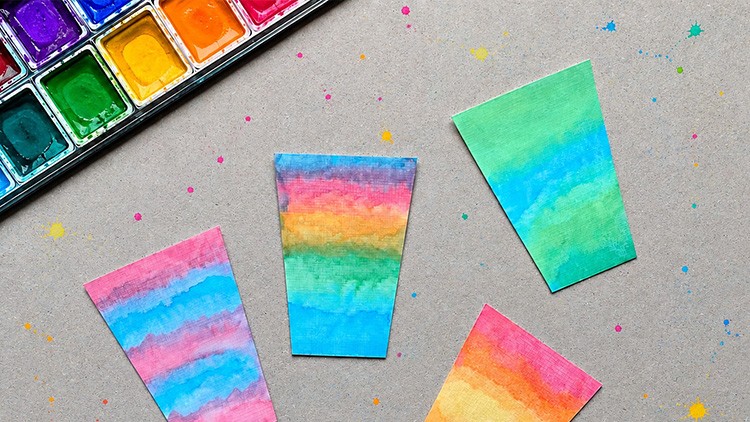 Vier bemalte Papierstücke in Regenbogenfarben liegen auf grauem Hintergrund, daneben ein offener Wassermalkasten.