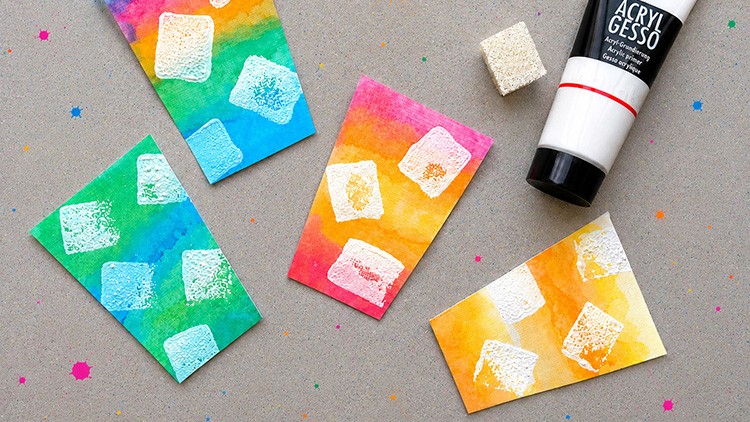 Bunte Karten mit weißem Schwammmuster auf grauem Untergrund, daneben ein weißer Schwamm und eine Tube Acrylgesso.