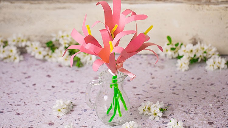 Eine durchsichtige Glasvase mit rosafarbenen Papierblumen und grünen Stielen steht auf hellem Untergrund mit weißen Blüten.