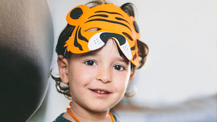 Kind trägt eine orangefarbene Tigermaske mit schwarzen Streifen und steht vor einem verschwommenen Hintergrund.