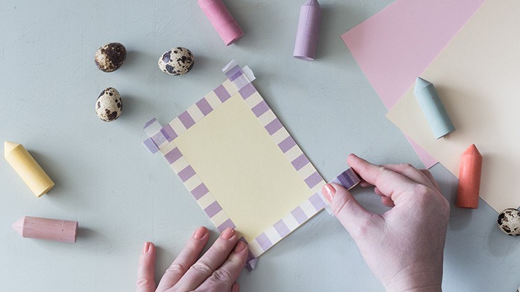 Zwei Hände dekorieren eine Karte mit lila-gestreiftem Klebeband, umgeben von Wachsmalstiften, buntem Papier und Wachteleiern.