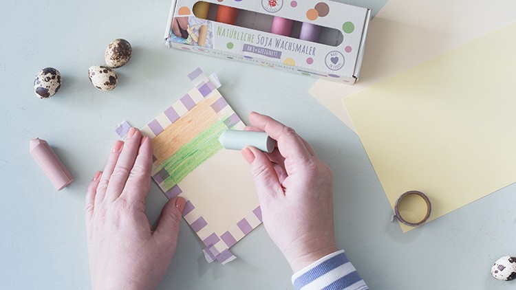 Hände bemalen Papier mit pastellgrünem Wachsmaler, umgeben von Kreppband, Wachsmalstiften, Wachteleiern und Papier.